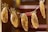 Several banana slices are hanging on a line using unique flower-shaped clips. The bananas appear to be dried, with a golden-brown color and visible dark spots along the length. The background is softly blurred, adding focus to the hanging bananas.