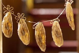 Several banana slices are hanging on a line using unique flower-shaped clips. The bananas appear to be dried, with a golden-brown color and visible dark spots along the length. The background is softly blurred, adding focus to the hanging bananas.
