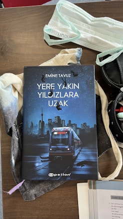 A book titled 'Yere Yakın Yıldızlara Uzak' by Emine Tavuz is placed on a brown wooden surface. The book cover features an illustration of a city skyline at night with a train in the foreground. A face mask and some fabric items are placed nearby, along with a partially visible stack of papers or a book on the right.