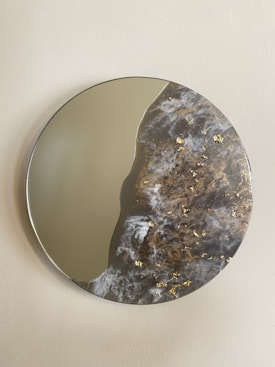 A round decorative mirror with an elegant design is displayed against a neutral-colored wall. The mirror is partially covered with an abstract pattern resembling ocean waves with swirling textures in grey, white, and brown, accented with gold leaf detail.