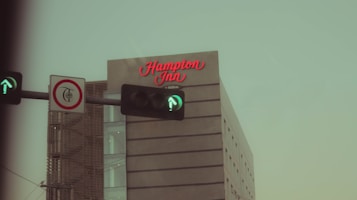 A modern hotel building with the signage 'Hampton Inn' in red cursive letters on its facade. In the foreground, a traffic light shows a green arrow, and there is a no left turn sign. The sky appears overcast, contributing to a somewhat muted atmosphere.