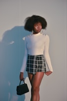 A person stands against a plain background, wearing a long-sleeved white top with metallic button details on the shoulders and a black and white plaid skirt. Large hoop earrings complement the outfit, and a small black quilted handbag is held in one hand. The individual has an afro hairstyle and displays a thoughtful expression.