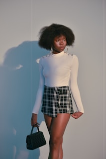 A person stands against a plain background, wearing a long-sleeved white top with metallic button details on the shoulders and a black and white plaid skirt. Large hoop earrings complement the outfit, and a small black quilted handbag is held in one hand. The individual has an afro hairstyle and displays a thoughtful expression.