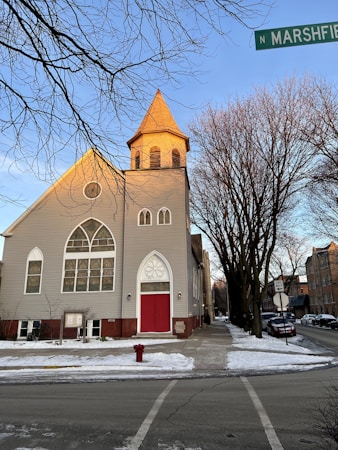 A church with light gray siding and a red door stands at the corner of a street. The architecture includes large arched windows with decorative designs. Bare trees line the sidewalk, and there is snow on the ground. A street sign indicates the location as N Marshfield.