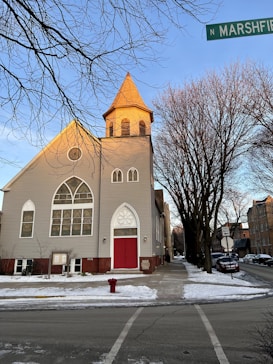 A church with light gray siding and a red door stands at the corner of a street. The architecture includes large arched windows with decorative designs. Bare trees line the sidewalk, and there is snow on the ground. A street sign indicates the location as N Marshfield.