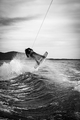A person is performing an aerial trick while wakeboarding on a body of water. The image is in black and white, capturing the dynamic movement of the wakeboarder amidst splashing waves. The background features a hilly landscape under a cloudy sky.