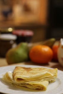 A folded crepe sits on a white plate in the foreground, displaying a golden-brown color and light, fluffy texture. In the blurred background, there are assorted fruits including an orange and a pear, along with jars that appear to contain spreads.