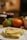 A folded crepe sits on a white plate in the foreground, displaying a golden-brown color and light, fluffy texture. In the blurred background, there are assorted fruits including an orange and a pear, along with jars that appear to contain spreads.