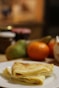 A folded crepe sits on a white plate in the foreground, displaying a golden-brown color and light, fluffy texture. In the blurred background, there are assorted fruits including an orange and a pear, along with jars that appear to contain spreads.