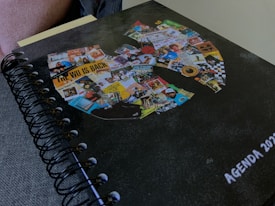 A spiral-bound agenda for the year 2023 with a cover that features a large collage of various images and text. The collage forms a recognizable symbol and includes pictures, posters, album covers, and text snippets in assorted colors.
