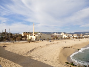 A sandy beach bordered by gentle ocean waves. In the background, there are industrial buildings with tall smokestacks, a large structure with the words 'ANIS DEL MONO', and various palm trees scattered along a walkway. A few people are strolling along the beach and the promenade, giving a sense of leisure and relaxation. The sky is partly cloudy with a light blue hue.