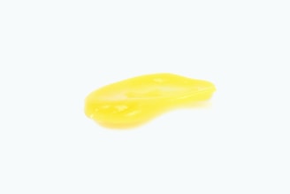 A small pool of bright yellow liquid with a glossy texture is set against a plain white background. The liquid appears smooth and slightly viscous.