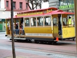 A vintage yellow and red San Francisco cable car is transporting several passengers. It bears the signage 'Powell & Mason Sts.' and 'Market Street Railway Co.' In the background, there are urban retail stores, and some people can be seen sitting on the sides of the cable car.