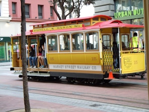 A vintage yellow and red San Francisco cable car is transporting several passengers. It bears the signage 'Powell & Mason Sts.' and 'Market Street Railway Co.' In the background, there are urban retail stores, and some people can be seen sitting on the sides of the cable car.