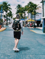 A person skateboards down a palm tree-lined street, wearing a backpack and casual clothing. The street is paved with blue tiles, and there are shops, bicycles, and people in the background enjoying what appears to be a sunny day.