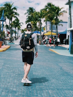 A person skateboards down a palm tree-lined street, wearing a backpack and casual clothing. The street is paved with blue tiles, and there are shops, bicycles, and people in the background enjoying what appears to be a sunny day.