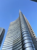 A modern skyscraper with a sleek, curved facade made primarily of glass and steel reaches into a clear blue sky. The building is part of a group of high-rise structures, showcasing contemporary architectural design with reflective surfaces.