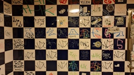 A tiled wall covered with a variety of graffiti and tags, featuring a mix of colors and styles. The tiles are arranged in a checkerboard pattern with alternating dark blue and white squares. Various tags and graffiti are in different colors such as red, blue, green, and black.