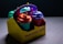 A brightly colored takeout box containing four cups with distinctive face-shaped lids. The box has a playful design with the text 'Shake & Shot To Go' on the side. Each cup is topped with a vibrant lid and straw, showcasing a whimsical approach to packaging.