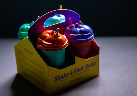 A brightly colored takeout box containing four cups with distinctive face-shaped lids. The box has a playful design with the text 'Shake & Shot To Go' on the side. Each cup is topped with a vibrant lid and straw, showcasing a whimsical approach to packaging.