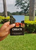 A hand is holding a notebook with the word 'CREATE' on the cover. The notebook has a colorful illustration featuring trees and a building on it. The background features a lush green garden with palm trees and flowering shrubs under an overcast sky.
