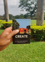 A hand is holding a notebook with the word 'CREATE' on the cover. The notebook has a colorful illustration featuring trees and a building on it. The background features a lush green garden with palm trees and flowering shrubs under an overcast sky.