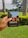 A hand is holding a notebook with the word 'CREATE' on the cover. The notebook has a colorful illustration featuring trees and a building on it. The background features a lush green garden with palm trees and flowering shrubs under an overcast sky.