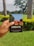 A hand is holding a notebook with the word 'CREATE' on the cover. The notebook has a colorful illustration featuring trees and a building on it. The background features a lush green garden with palm trees and flowering shrubs under an overcast sky.