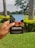 A hand is holding a notebook with the word 'CREATE' on the cover. The notebook has a colorful illustration featuring trees and a building on it. The background features a lush green garden with palm trees and flowering shrubs under an overcast sky.