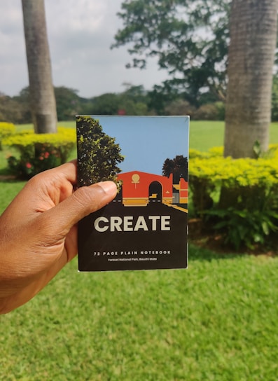 A hand is holding a notebook with the word 'CREATE' on the cover. The notebook has a colorful illustration featuring trees and a building on it. The background features a lush green garden with palm trees and flowering shrubs under an overcast sky.