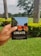 A hand is holding a notebook with the word 'CREATE' on the cover. The notebook has a colorful illustration featuring trees and a building on it. The background features a lush green garden with palm trees and flowering shrubs under an overcast sky.