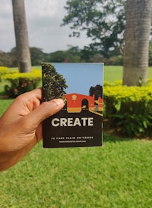 A hand is holding a notebook with the word 'CREATE' on the cover. The notebook has a colorful illustration featuring trees and a building on it. The background features a lush green garden with palm trees and flowering shrubs under an overcast sky.