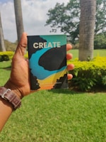 A hand is holding a colorful notebook with the word 'CREATE' printed on the cover. The background features a lush green garden with trees, bushes, and a partly cloudy sky.