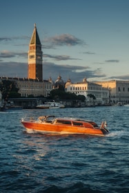A historic waterfront scene with a prominent tall clock tower under a slightly cloudy sky at dusk. In front, a stylish wooden boat is cruising through gentle waves on a large body of water. Surrounding the tower are a variety of classic and ornate buildings.