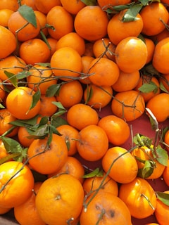 A large pile of fresh oranges with vibrant orange skin, scattered randomly. Some of the oranges are adorned with green leaves and thin stems, indicating they were recently picked from a tree. The lighting highlights their textures and colors, emphasizing the freshness of the fruit.