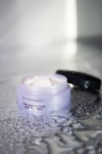 A jar of MAC Studio Moisture Cream is seen with its lid off, placed on a textured and wet reflective surface. The creamy substance inside is visible and appears smooth and ready for application, with some of the product spread on the surface around the jar.