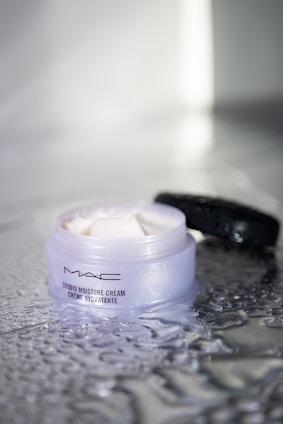 A jar of MAC Studio Moisture Cream is seen with its lid off, placed on a textured and wet reflective surface. The creamy substance inside is visible and appears smooth and ready for application, with some of the product spread on the surface around the jar.