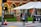 A person wearing a red cap and blue jacket is standing on a sidewalk near the University of Southern California entrance. The individual is looking down at their phone. The scene includes a brick building facade with large arched windows. A large white tent is set up nearby, with red umbrellas providing a splash of color. A traffic signal displays a hand for 'stop,' and a traffic cone is placed near the entrance.