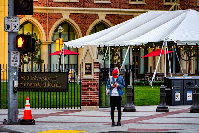 A person wearing a red cap and blue jacket is standing on a sidewalk near the University of Southern California entrance. The individual is looking down at their phone. The scene includes a brick building facade with large arched windows. A large white tent is set up nearby, with red umbrellas providing a splash of color. A traffic signal displays a hand for 'stop,' and a traffic cone is placed near the entrance.