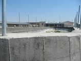 A concrete structure with several vertical poles rising from it is set under a clear blue sky. In the background, there are industrial-style buildings with flat roofs and a few windows. The surface of the concrete appears weathered and textured.