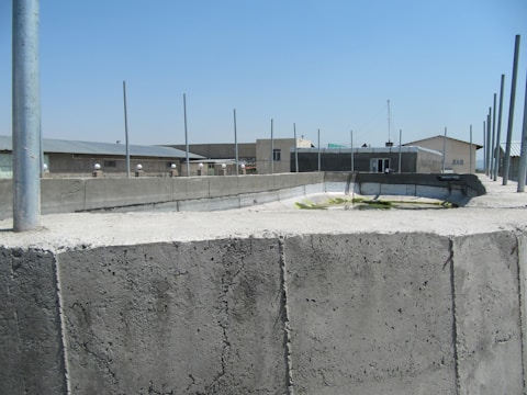 A concrete structure with several vertical poles rising from it is set under a clear blue sky. In the background, there are industrial-style buildings with flat roofs and a few windows. The surface of the concrete appears weathered and textured.