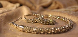 A close-up view of a sparkling jewelry piece placed on a softly textured fabric surface. The jewelry appears to be encrusted with small, shiny gemstones and has a luxurious, elegant appearance.