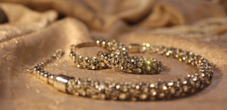 A close-up view of a sparkling jewelry piece placed on a softly textured fabric surface. The jewelry appears to be encrusted with small, shiny gemstones and has a luxurious, elegant appearance.