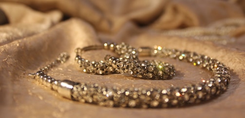 A close-up view of a sparkling jewelry piece placed on a softly textured fabric surface. The jewelry appears to be encrusted with small, shiny gemstones and has a luxurious, elegant appearance.