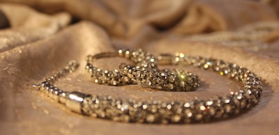 A close-up view of a sparkling jewelry piece placed on a softly textured fabric surface. The jewelry appears to be encrusted with small, shiny gemstones and has a luxurious, elegant appearance.