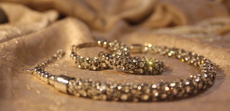 A close-up view of a sparkling jewelry piece placed on a softly textured fabric surface. The jewelry appears to be encrusted with small, shiny gemstones and has a luxurious, elegant appearance.