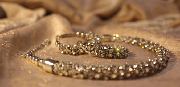 A close-up view of a sparkling jewelry piece placed on a softly textured fabric surface. The jewelry appears to be encrusted with small, shiny gemstones and has a luxurious, elegant appearance.