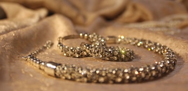 A close-up view of a sparkling jewelry piece placed on a softly textured fabric surface. The jewelry appears to be encrusted with small, shiny gemstones and has a luxurious, elegant appearance.