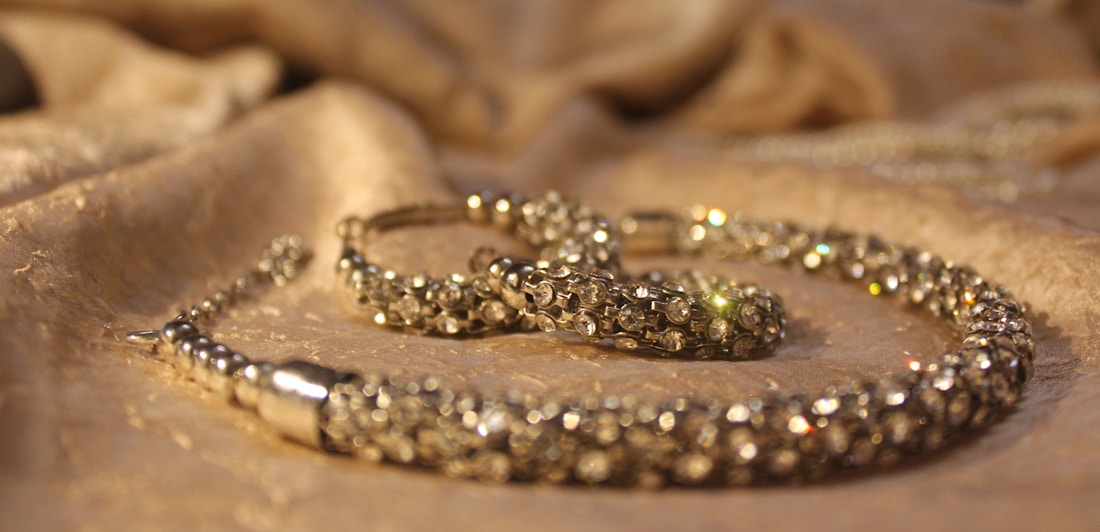 A close-up view of a sparkling jewelry piece placed on a softly textured fabric surface. The jewelry appears to be encrusted with small, shiny gemstones and has a luxurious, elegant appearance.