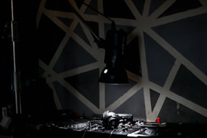 A dimly lit room with a spotlight hanging over a DJ mixer and headphones. The background features a geometric pattern with beige lines on a dark surface, creating an abstract look.